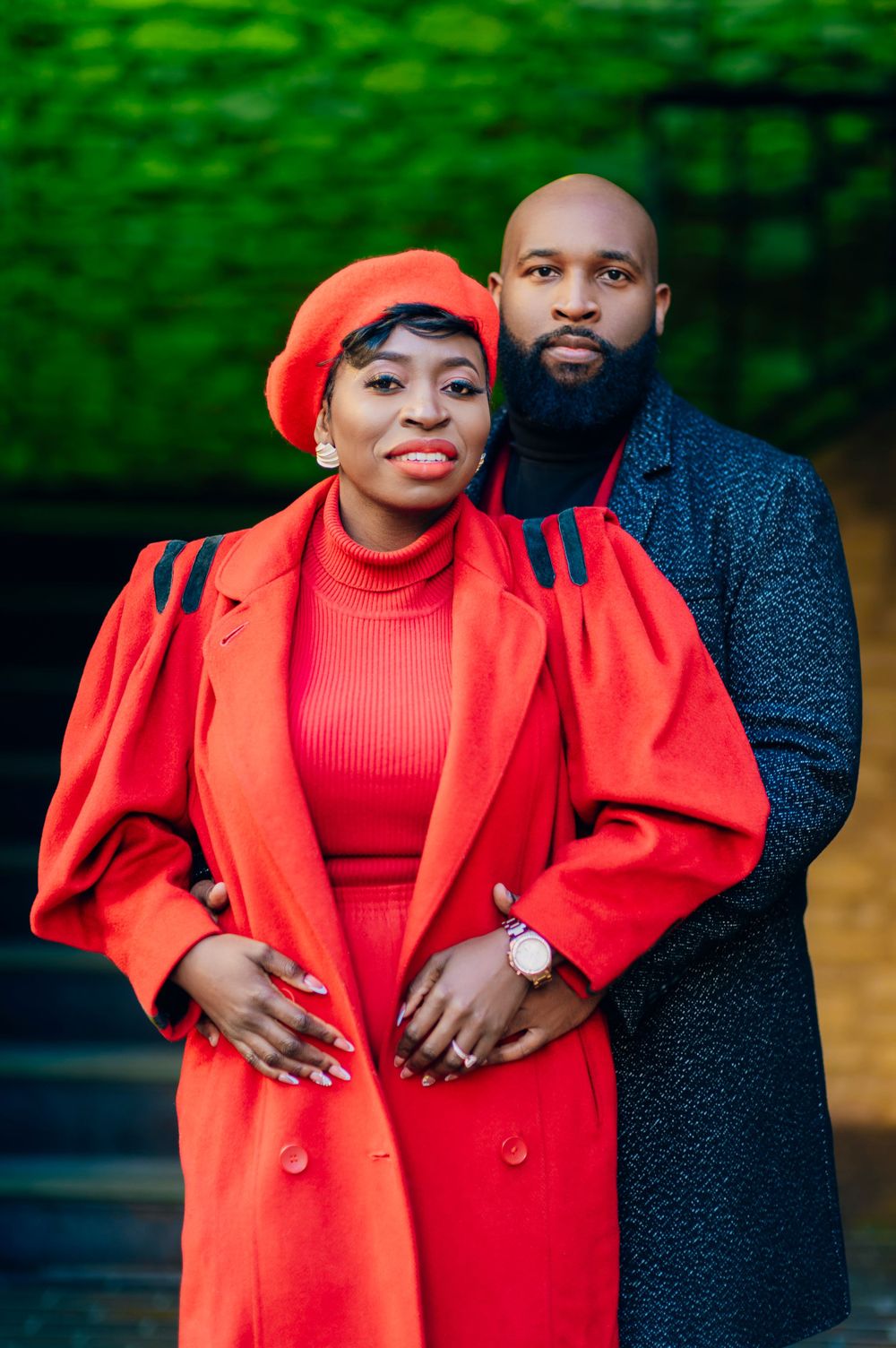A couple poses outdoors in coordinating outfits featuring a bright red coat and dark blue sweater against a green background.