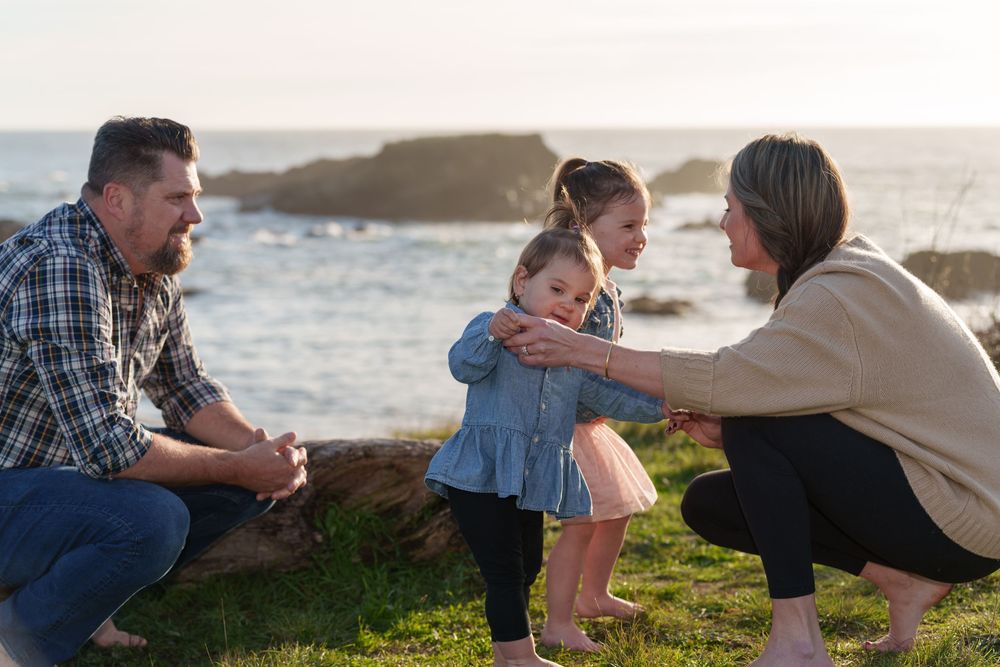 Family enjoying a playful moment together on grassy hillside overlooking the ocean at sunset.