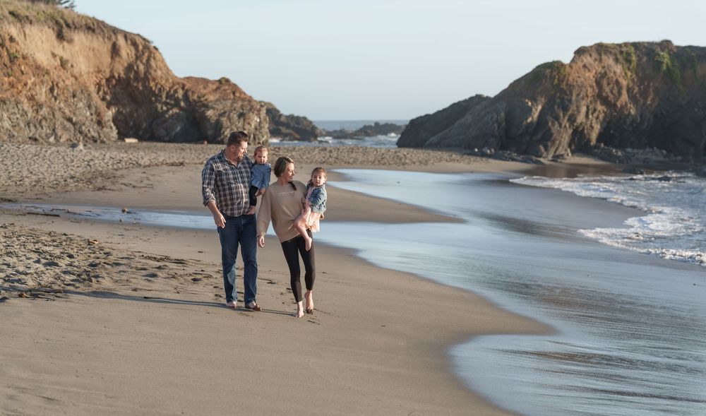 Couple walking along scenic Sea Ranch coastal beach with rocky cliffs and crashing waves in the background.