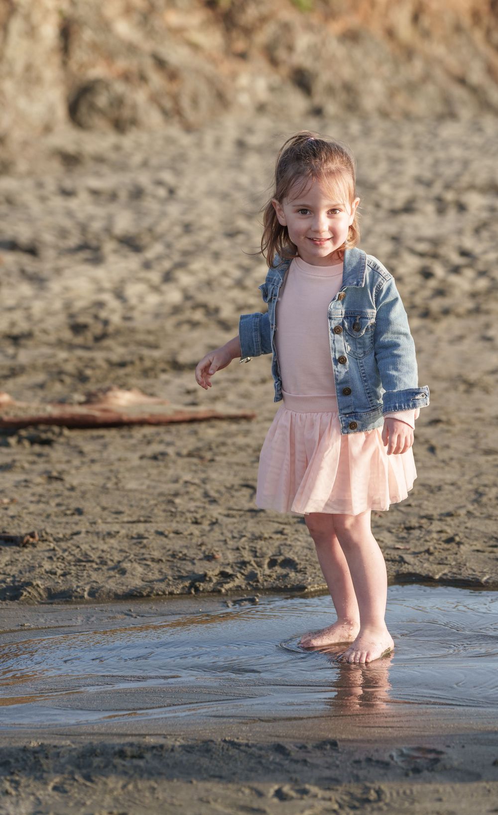 Young child in pink dress and denim jacket stands barefoot at the beach during golden hour.