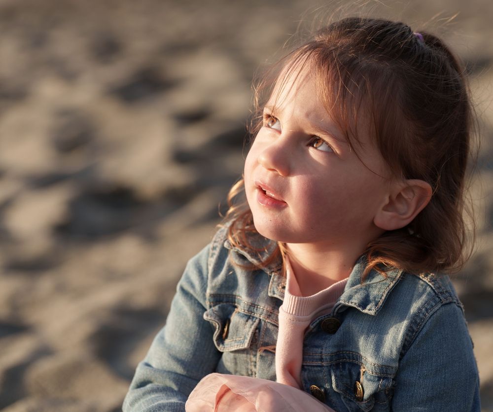 A young child in a denim jacket gazes upward with a dreamy expression during golden hour at the beach.