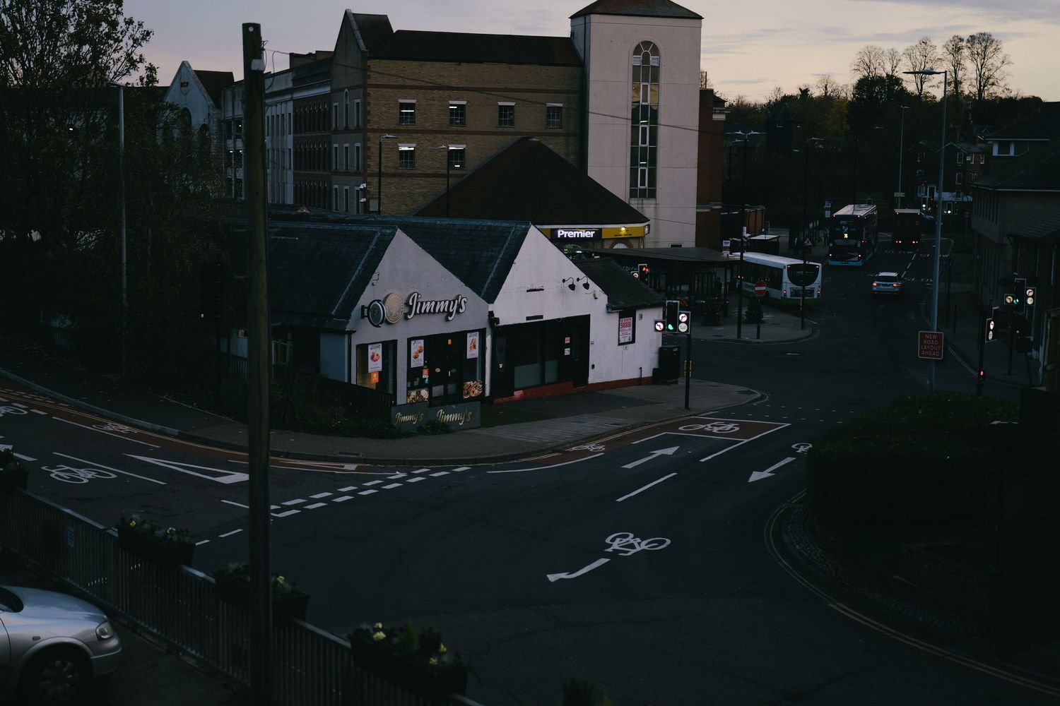 A dark street intersection at dusk with buildings and trees silhouetted against the dimming sky.