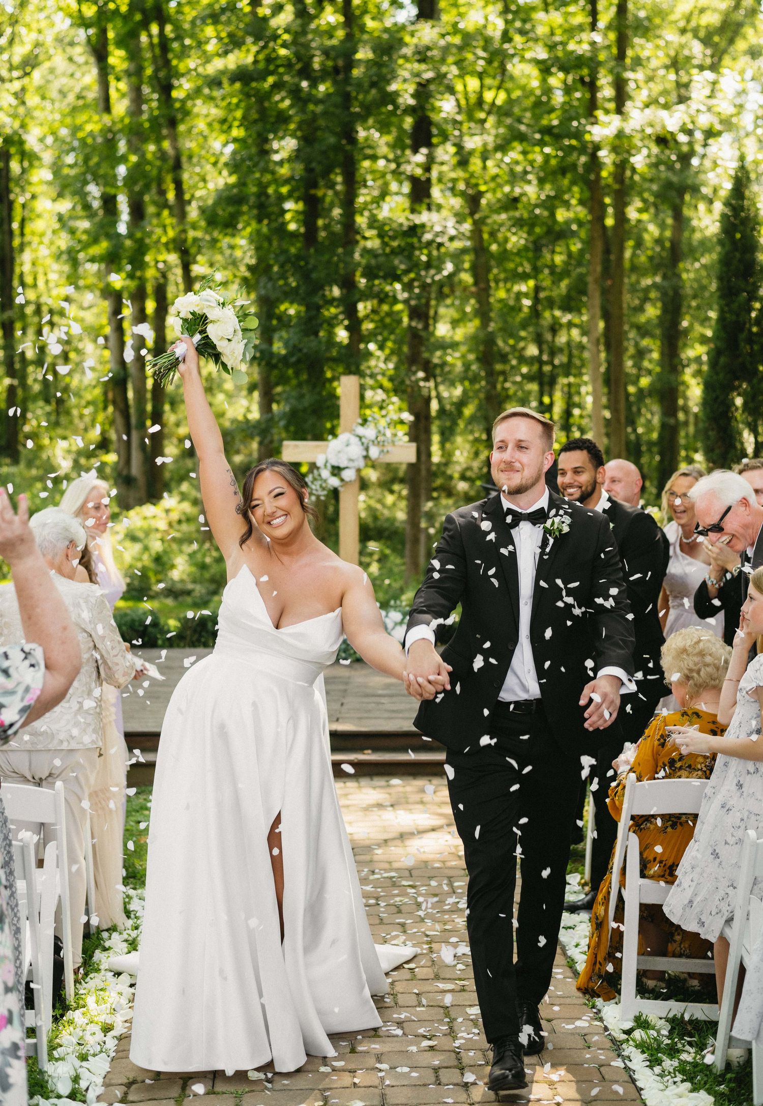 Newlyweds celebrate with confetti while walking down stone path at outdoor forest wedding ceremony.