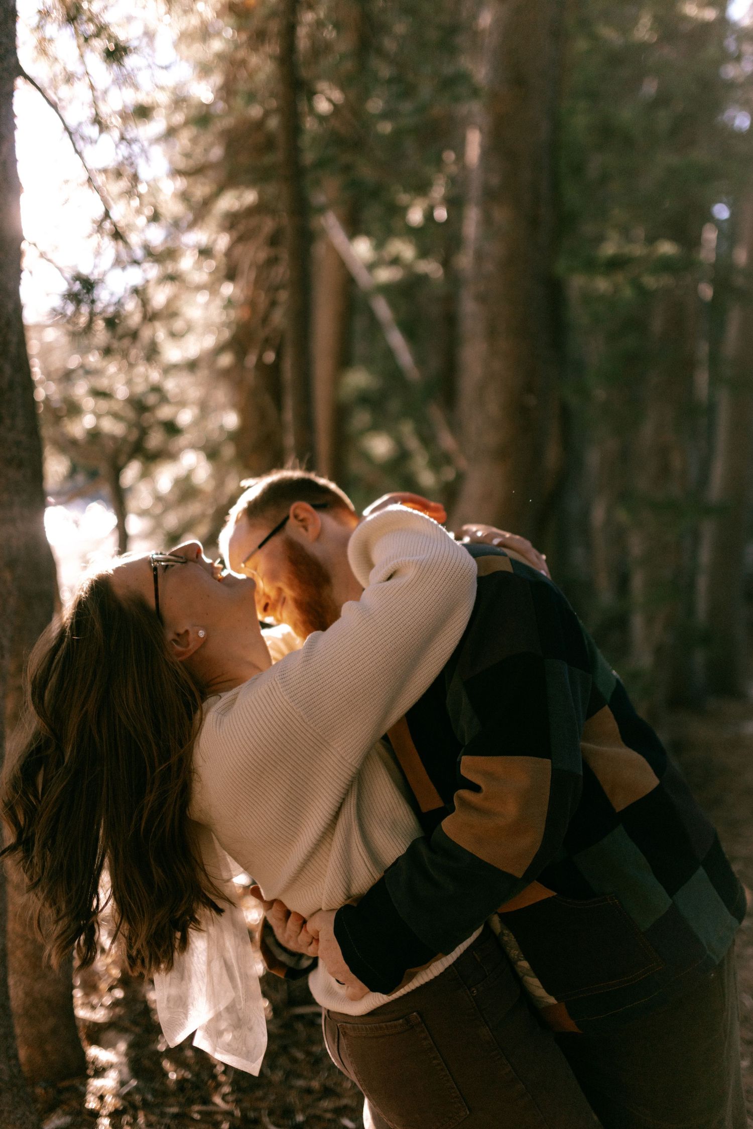 A romantic embrace between two people in a sunlit forest, backlit by warm golden light filtering through the trees.
