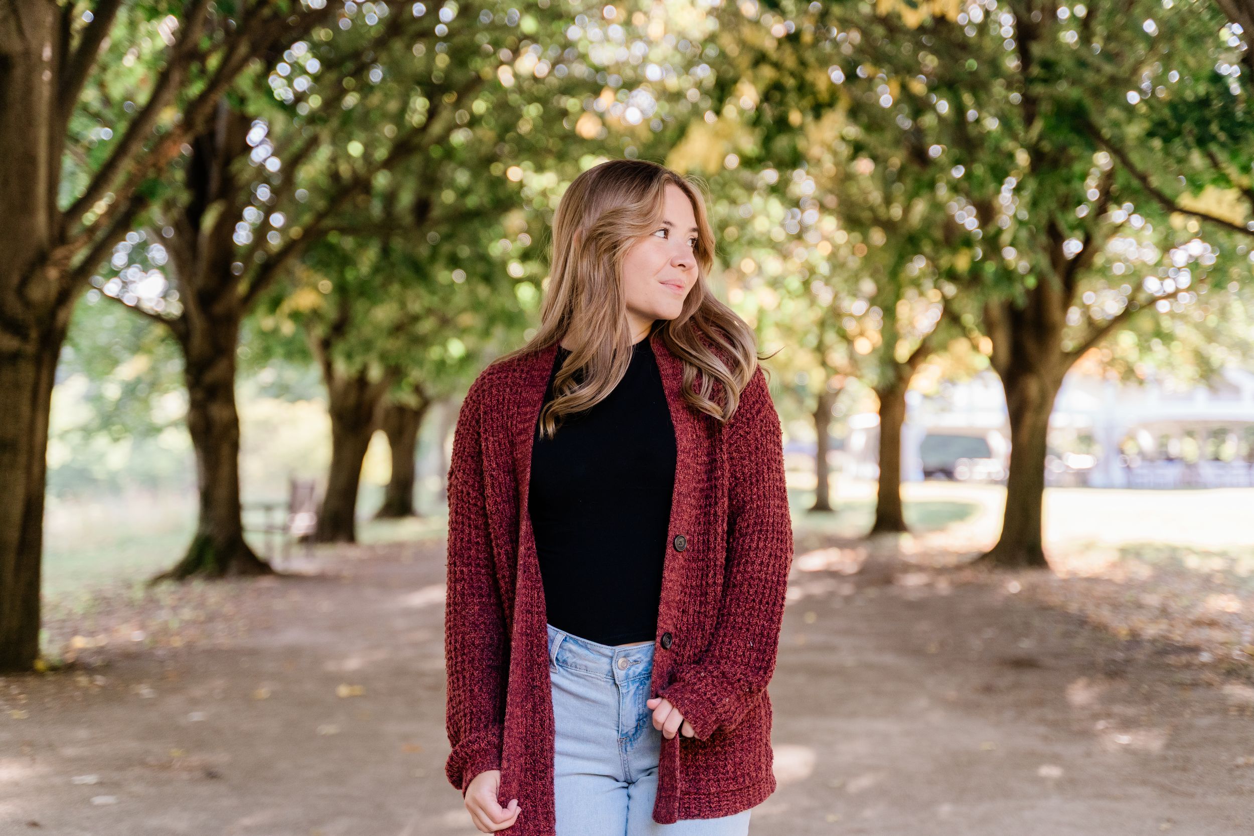 Woman in burgundy cardigan and jeans poses outdoors under tree-lined path in autumn setting.
