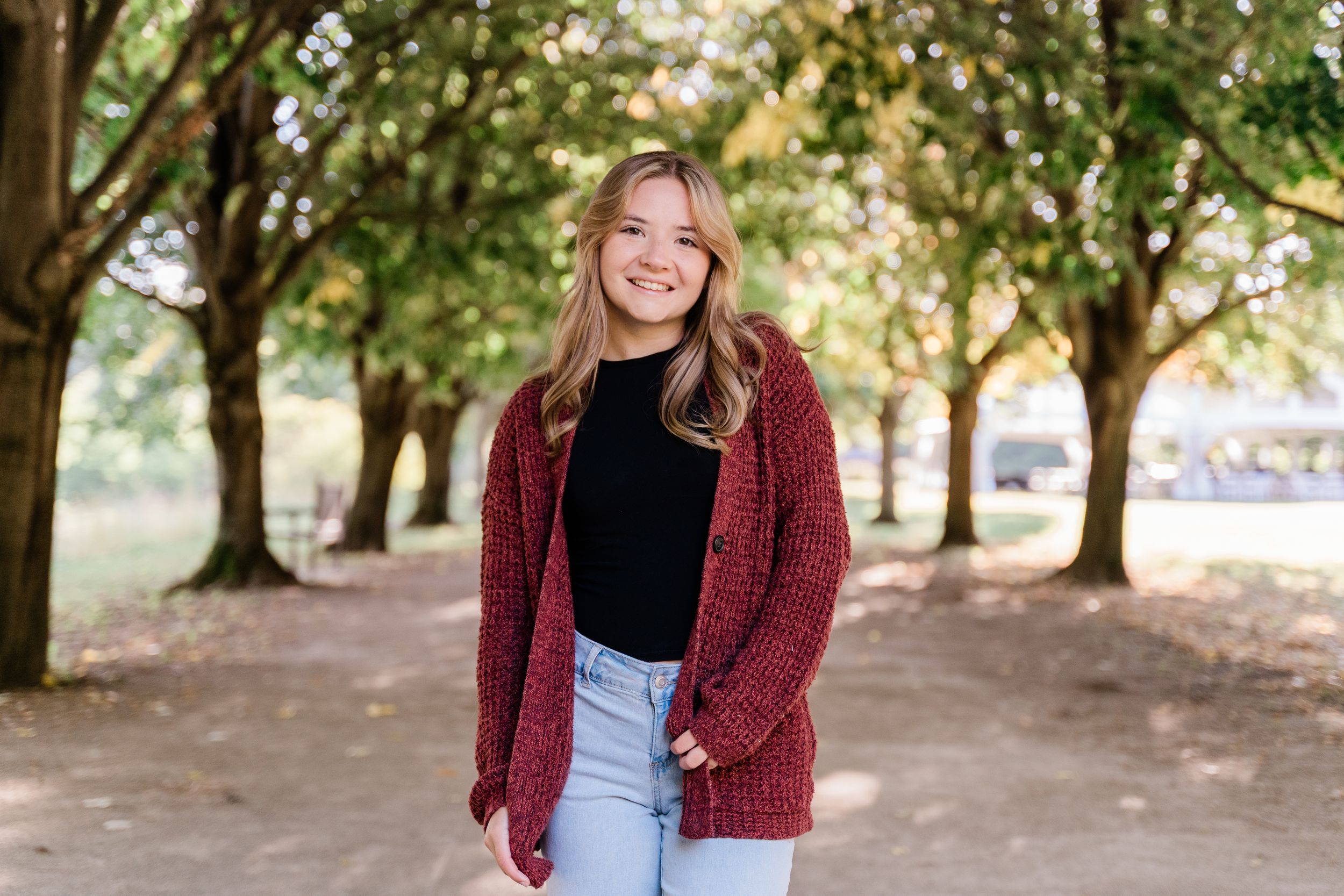 A person in a burgundy cardigan and jeans stands smiling on a tree-lined path during autumn.