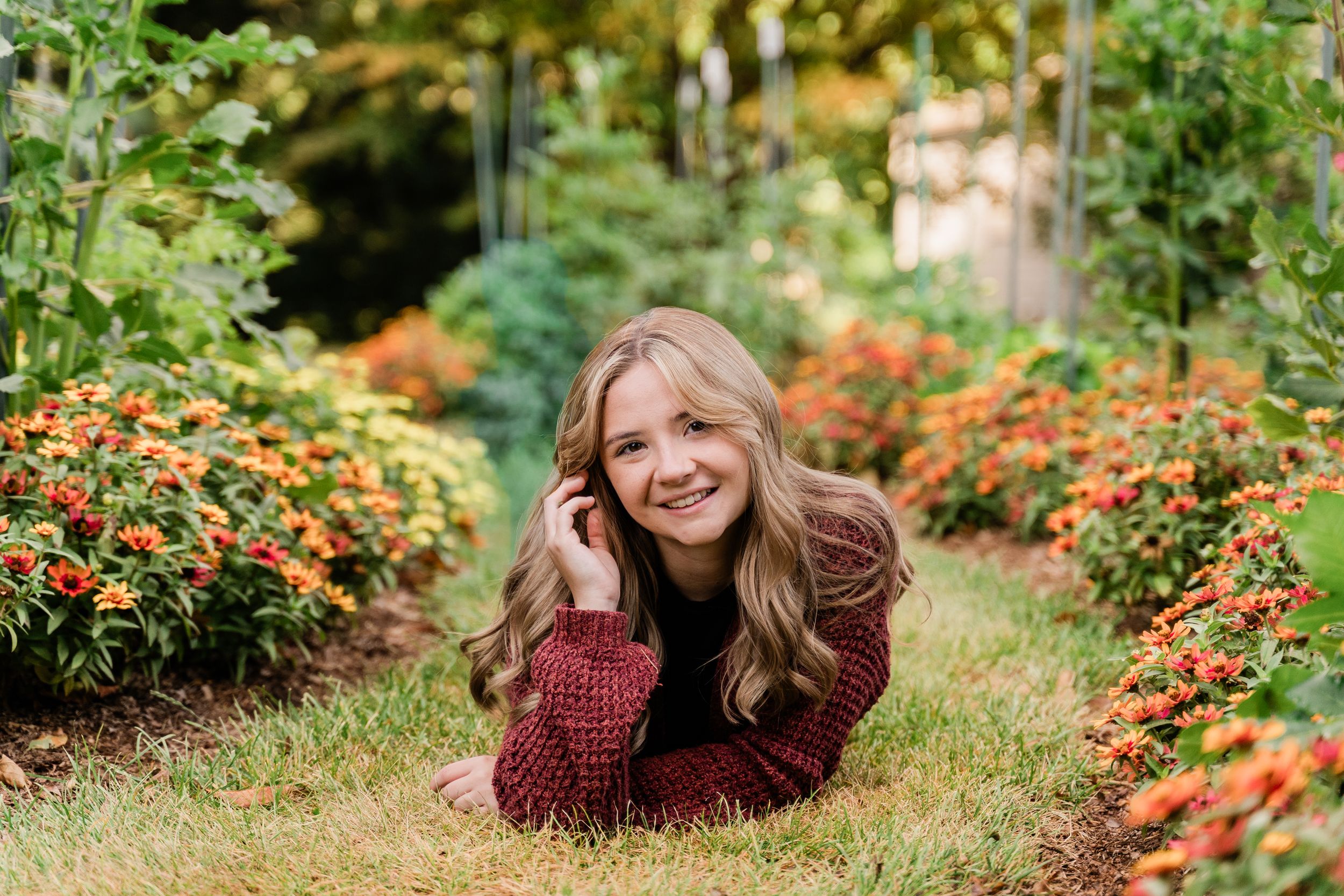 Someone lies on grass path in garden wearing burgundy sweater surrounded by vibrant orange lantana flowers in bloom.