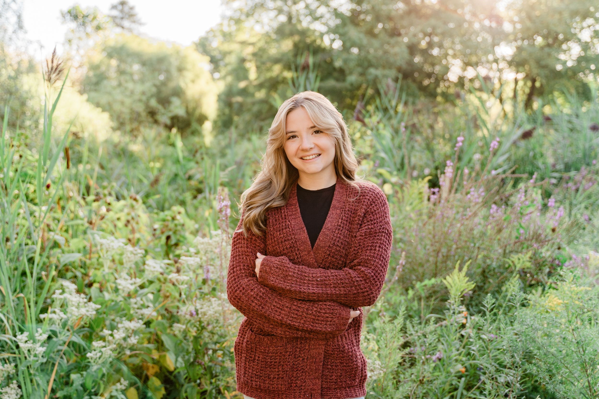 Person in a cozy rust-colored sweater smiles warmly while standing in a sunlit garden with tall greenery.