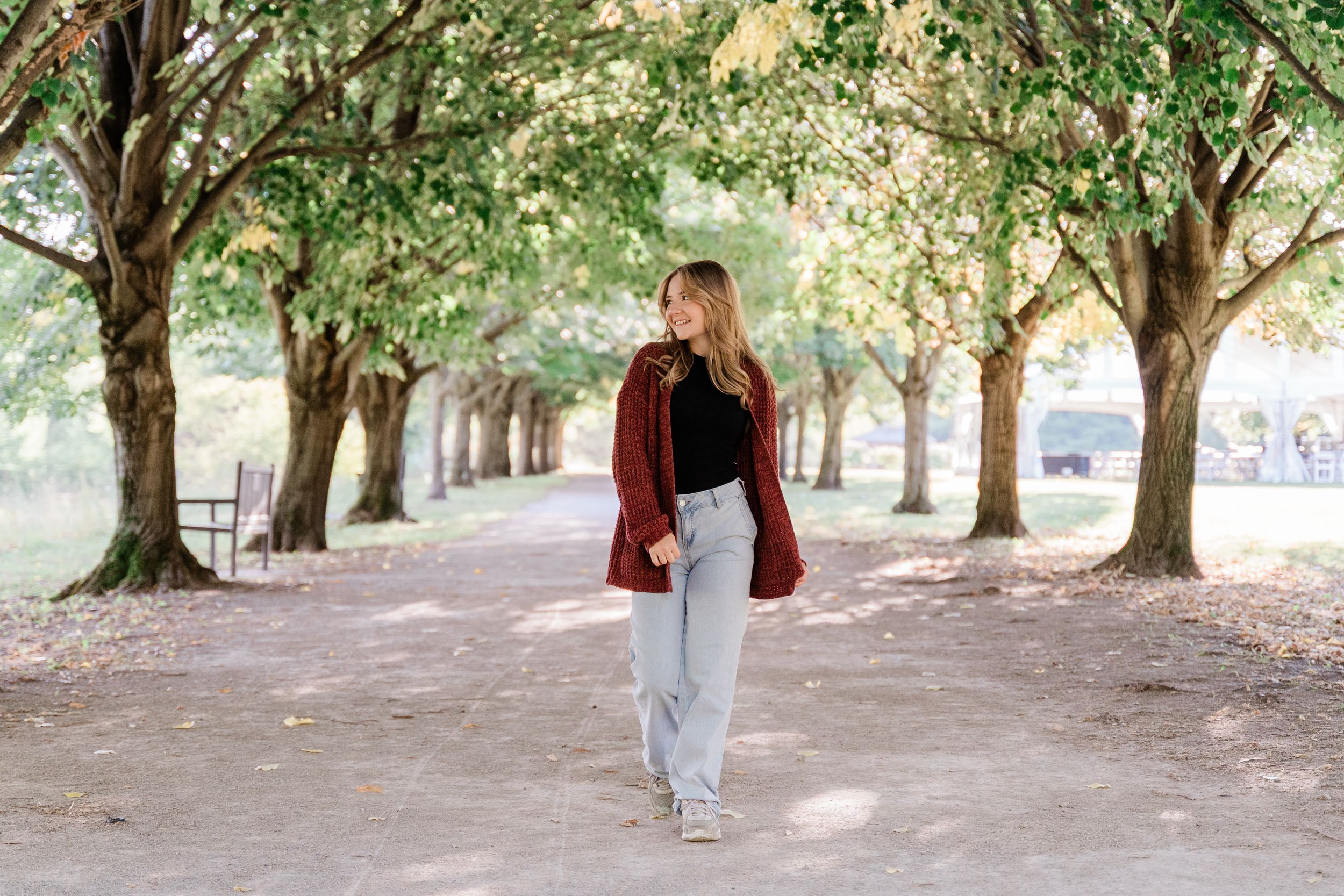 A person wearing a burgundy jacket and jeans walks through a tree-lined path on a sunny autumn day.