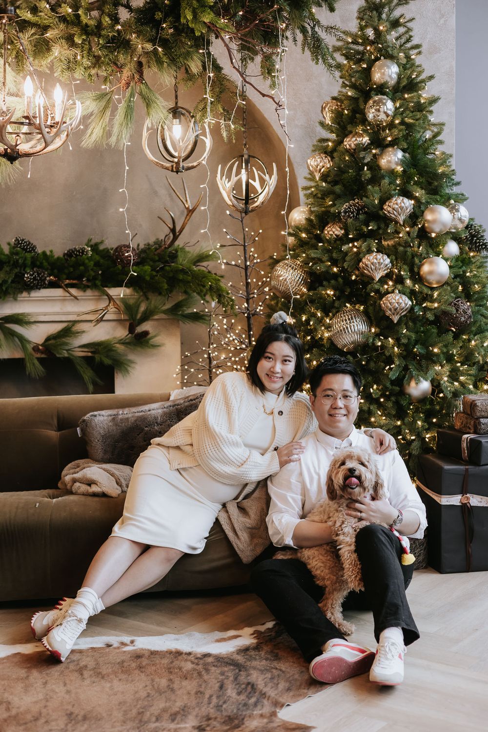 A cozy Christmas photoshoot featuring a couple in white sweaters with their small brown dog by a decorated tree.