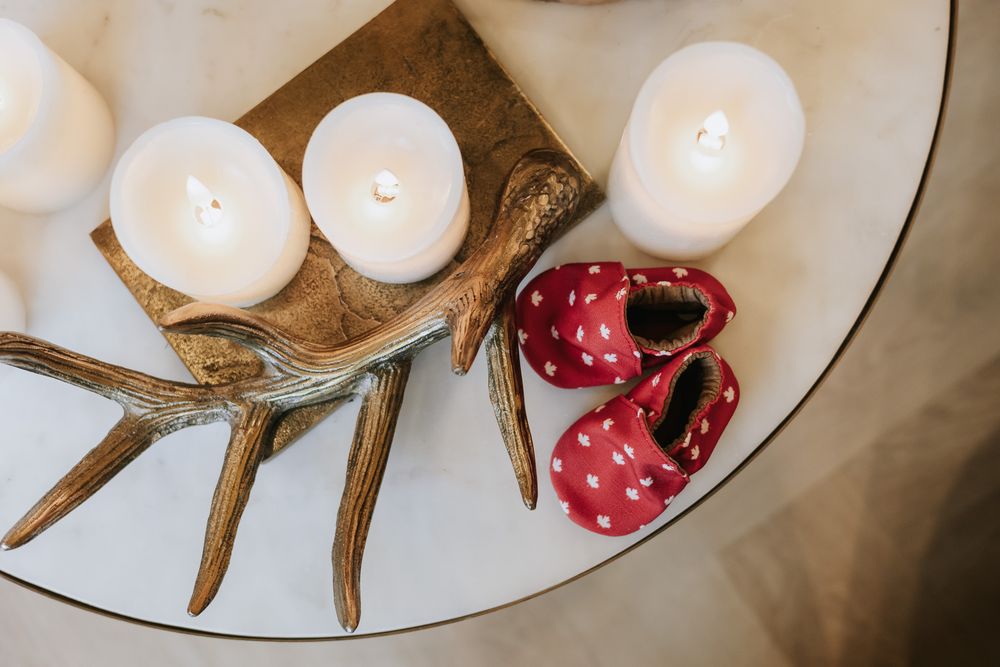 White candles on driftwood next to red polka dot baby shoes on a white surface.
