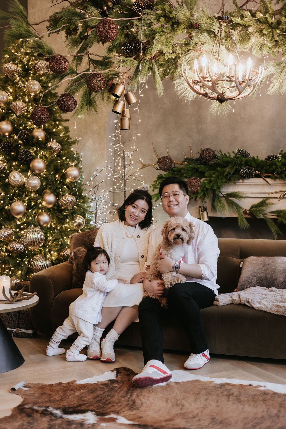A family wearing matching white outfits sits together on a brown couch under decorative greenery and twinkling lights.