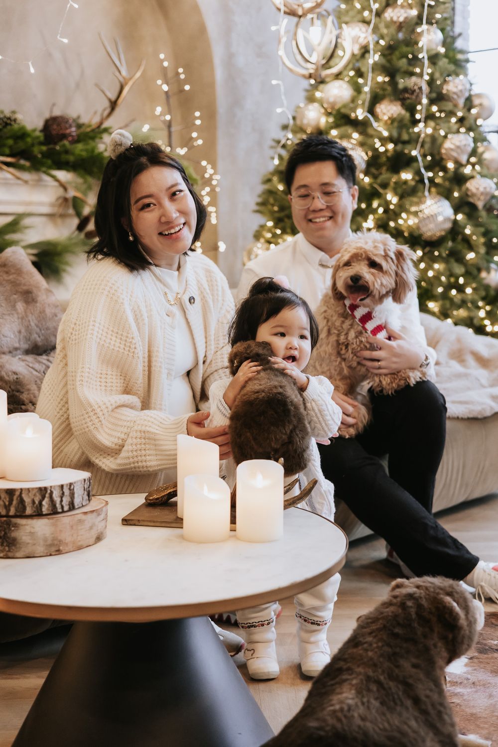 A cozy winter scene with people gathered around a white coffee table with lit candles and Christmas trees in the background.