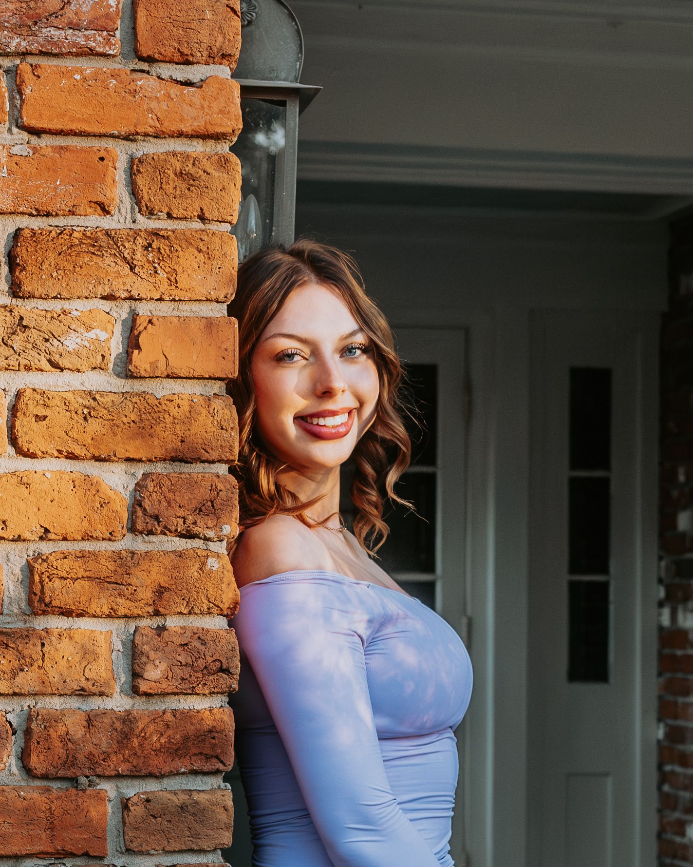 A smiling figure in a light blue off-shoulder dress poses against a rustic brick wall on a sunny day.