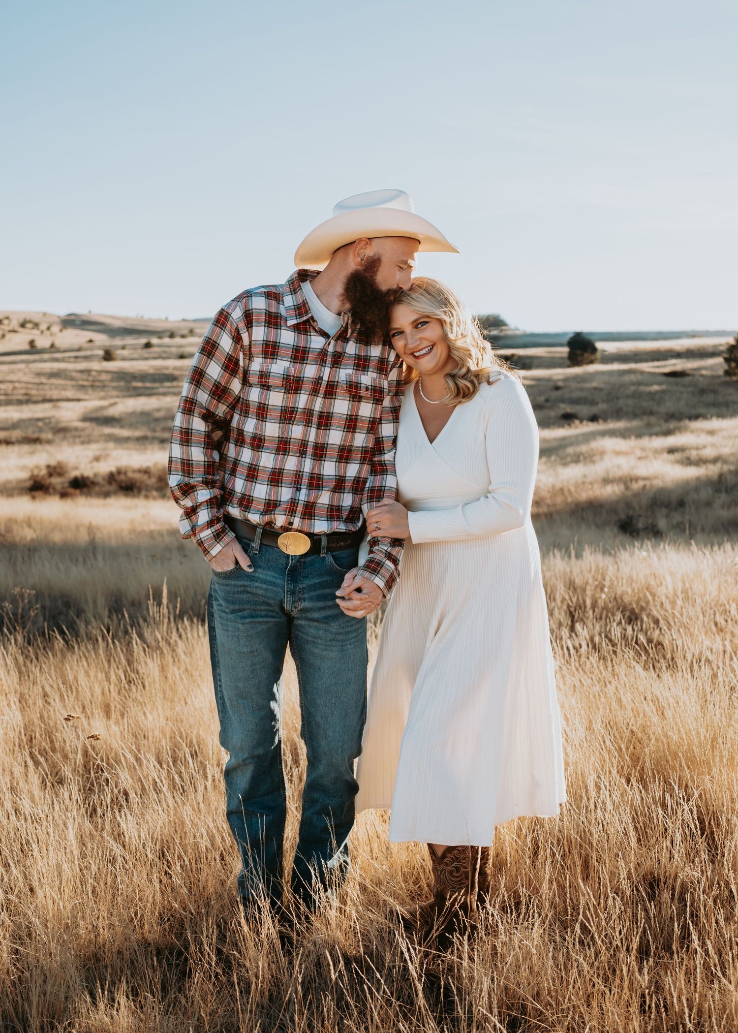 Romantic couple in western attire embraces in golden prairie grass during sunset photoshoot.
