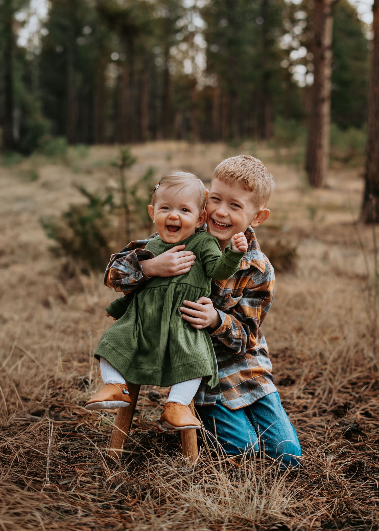 Two children laughing together in a pine forest setting during an autumn photoshoot.