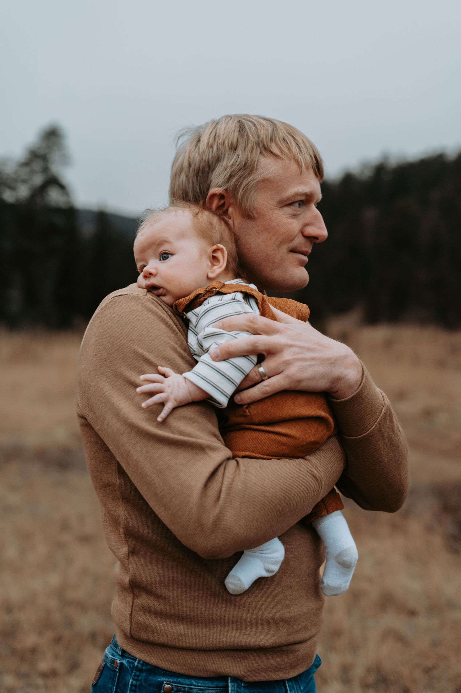 A parent in a brown sweater holds and embraces a baby in a white onesie against a moody outdoor backdrop.