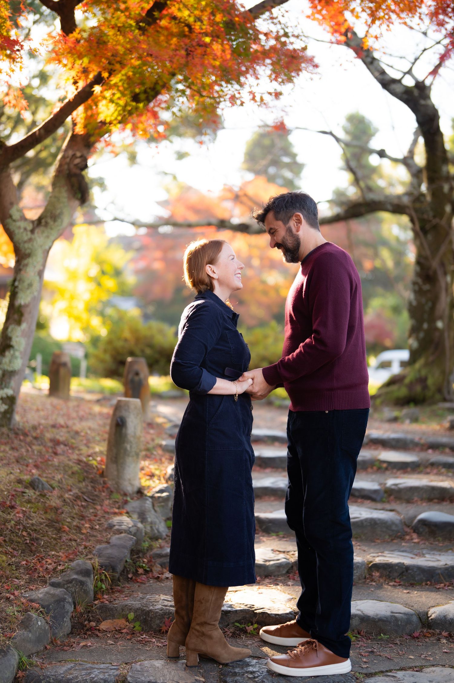 A couple holds hands and gazes at each other on stone steps surrounded by autumn foliage.