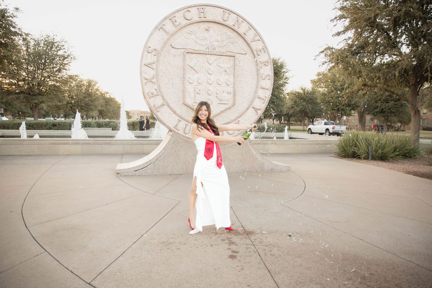 A graduate in a white dress poses in front of a large Texas Tech University seal on campus.