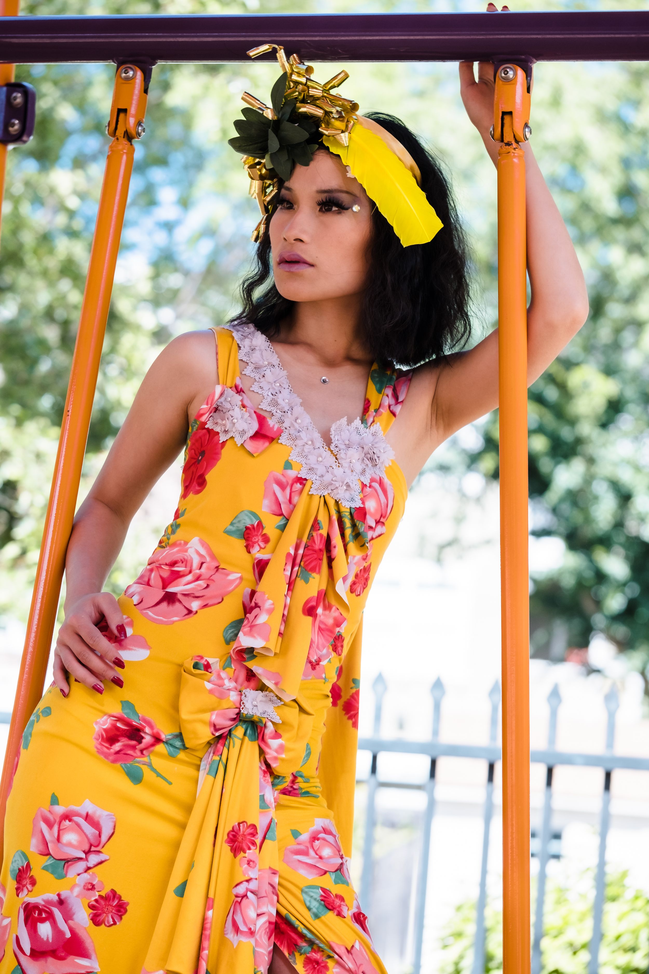 A person in a vibrant yellow floral dress poses outdoors near orange poles against a blurred natural background. Playground photoshoot