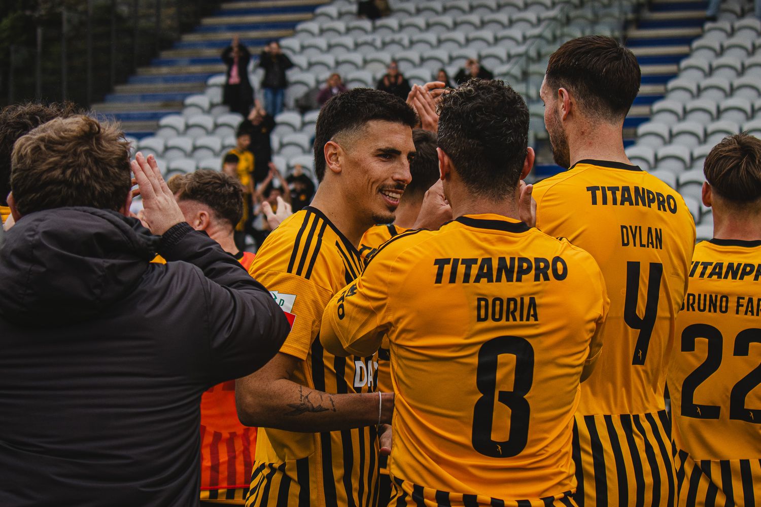 Team huddle of soccer players wearing yellow jerseys with TitanPro sponsorship on a cloudy day.