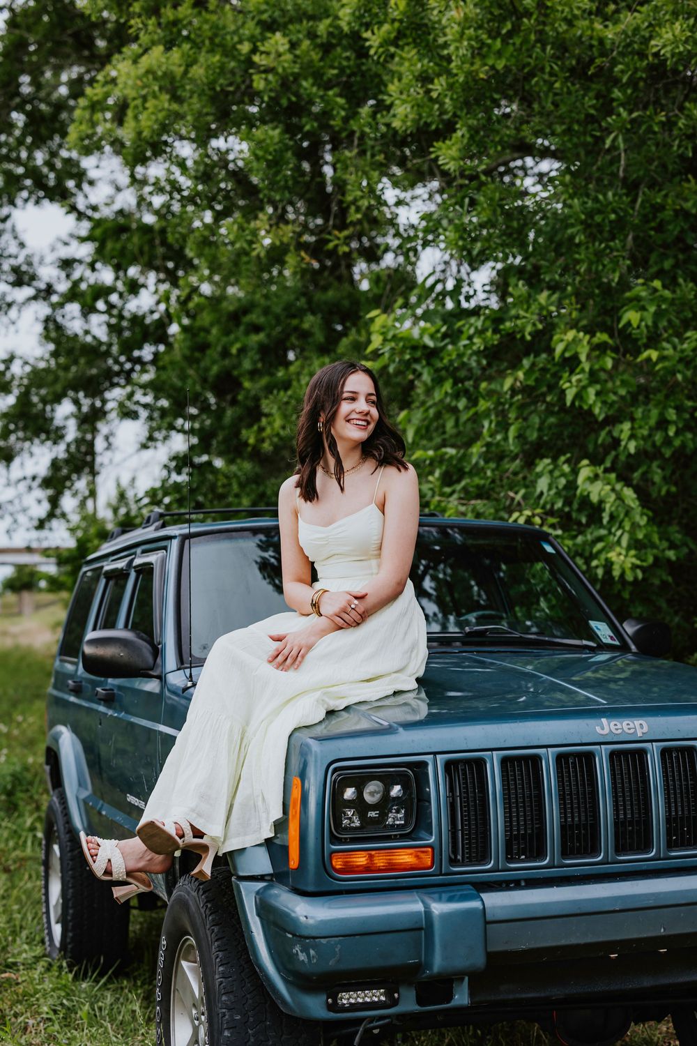 A white dress contrasts beautifully against a vintage blue Jeep in a lush green outdoor setting.