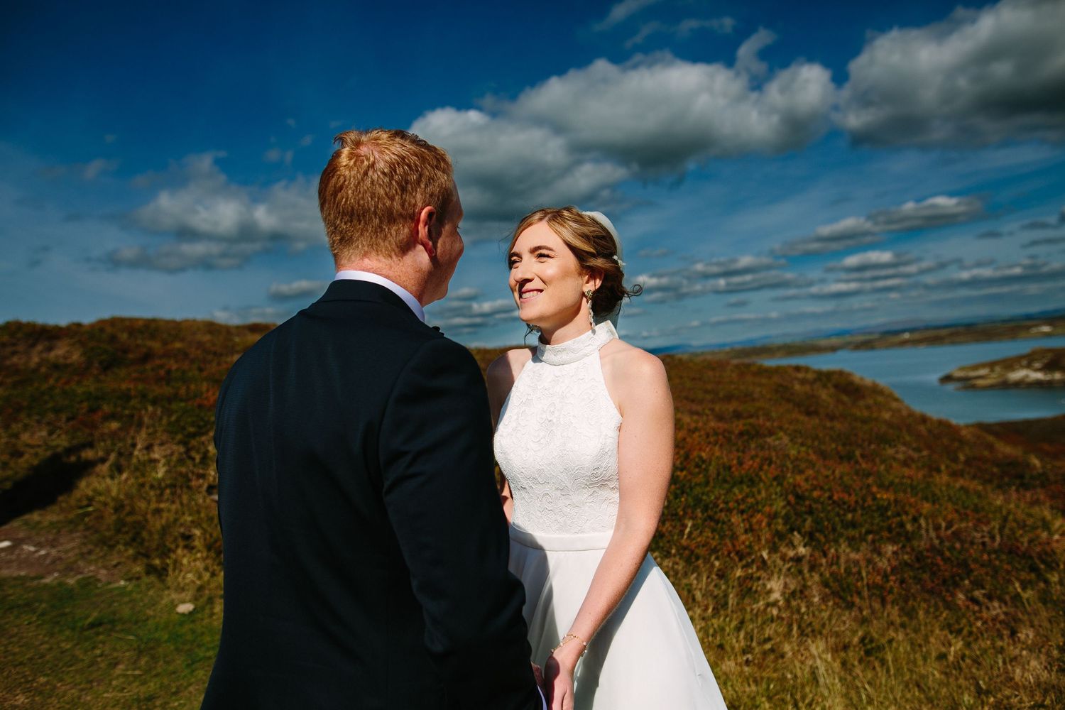 A couple in wedding attire stands together on a grassy hillside beneath a bright blue sky with white clouds.