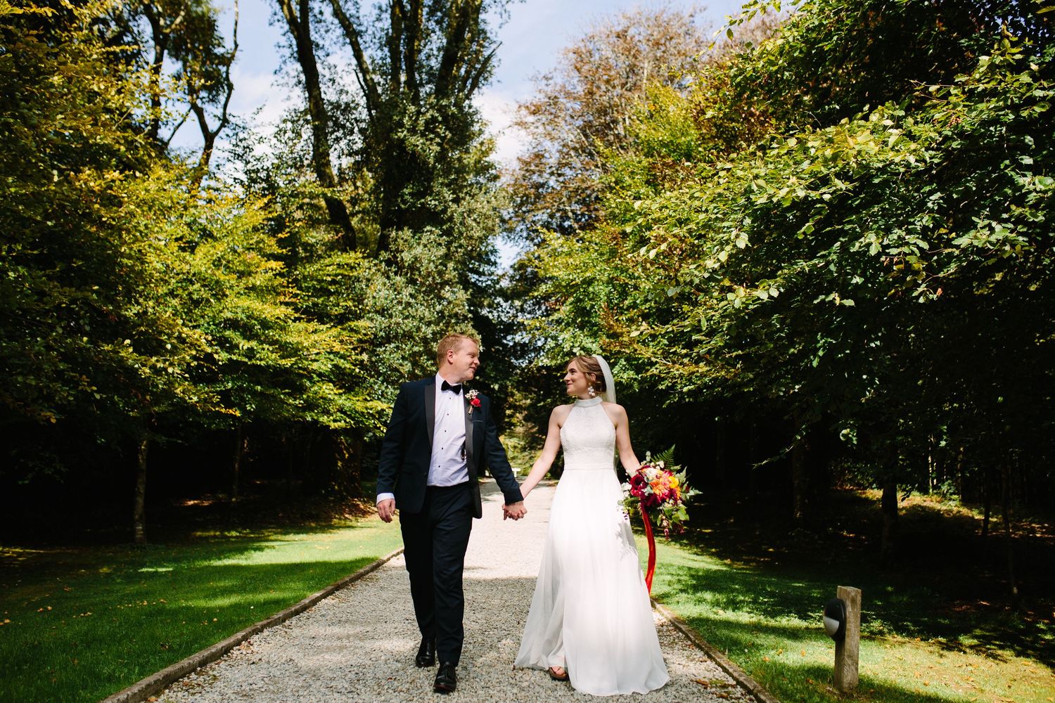 A bride and groom walk hand in hand down a tree-lined path surrounded by lush autumn foliage.