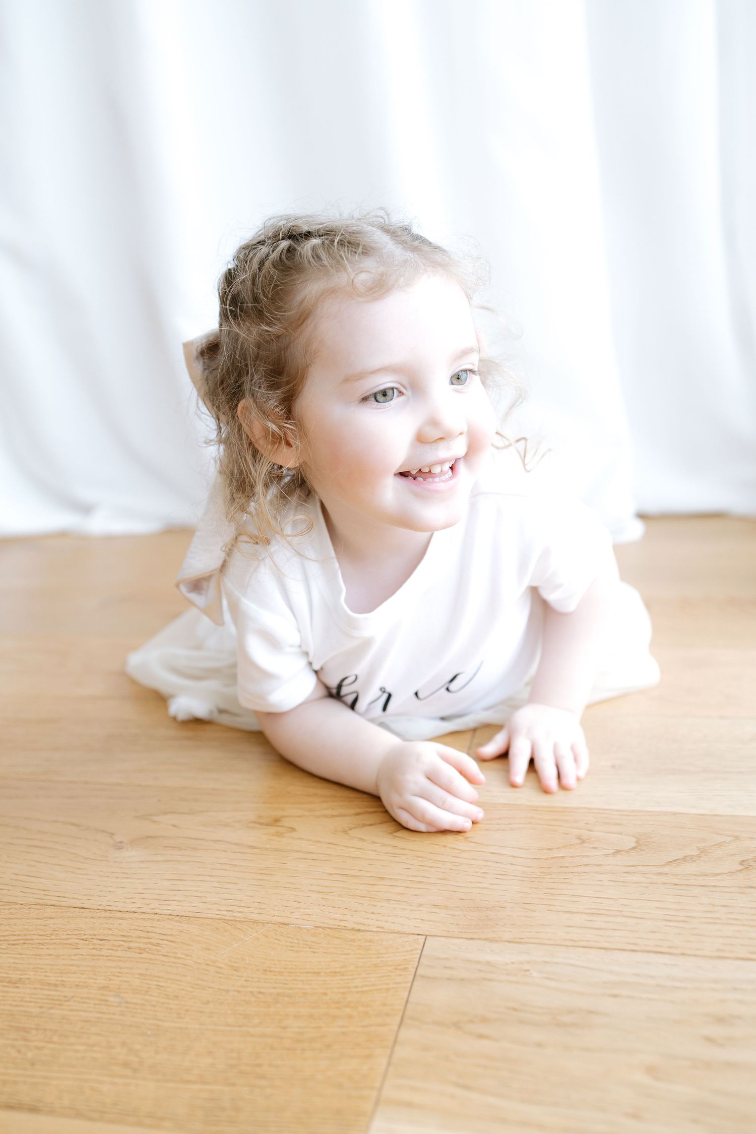 Baby in beige romper sits on white fluffy rug with pink flowers and green tropical leaves as decorative accents.