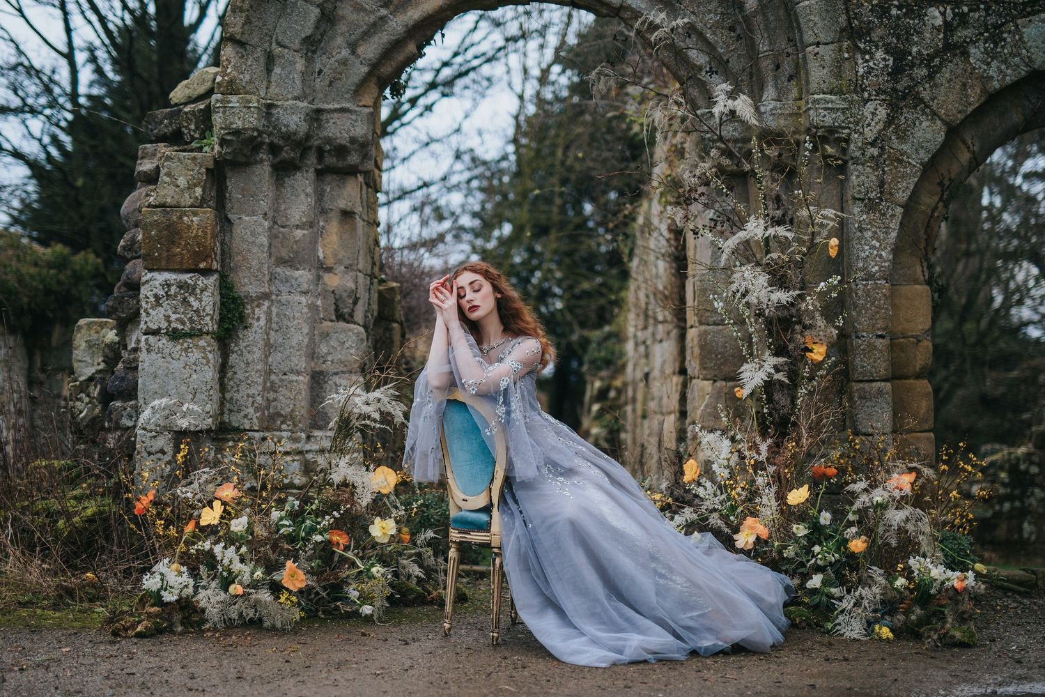An image of a woman in a purple fairy-style dress sat with floral decorations against stone archway ruins in a moody outdoor setting.