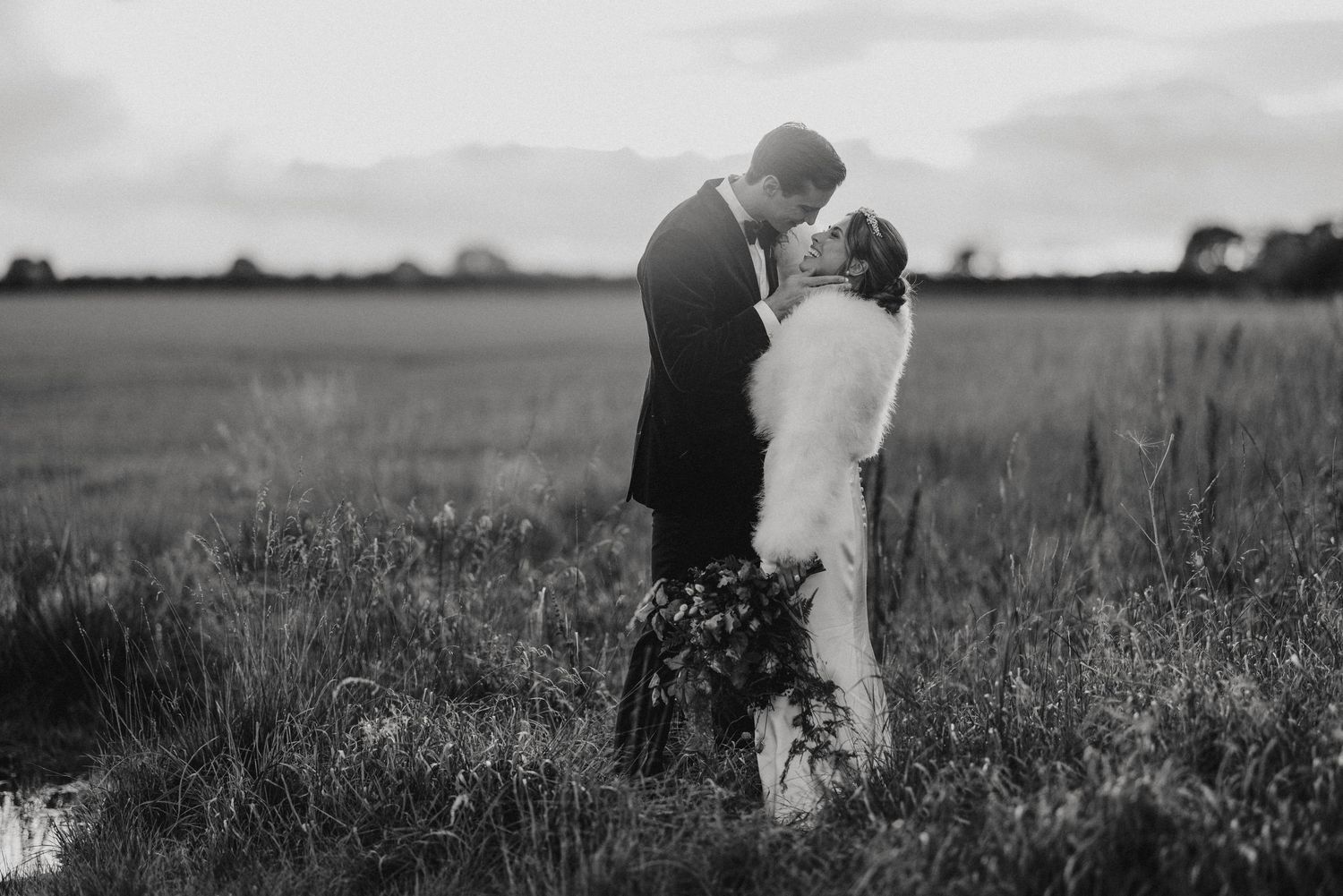 A couple shares a romantic embrace in a sun-drenched meadow filled with wildflowers and tall grass.