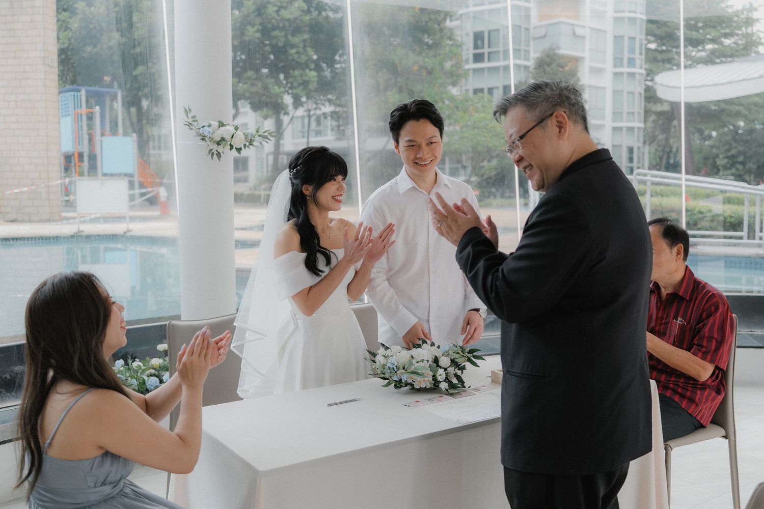 Wedding ceremony taking place in a bright modern space with white pillars and floral decorations.