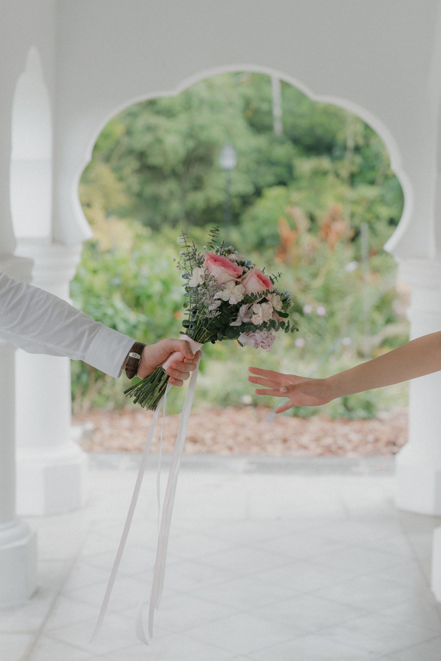 A delicate floral wedding bouquet being passed between hands in an arched white doorway.