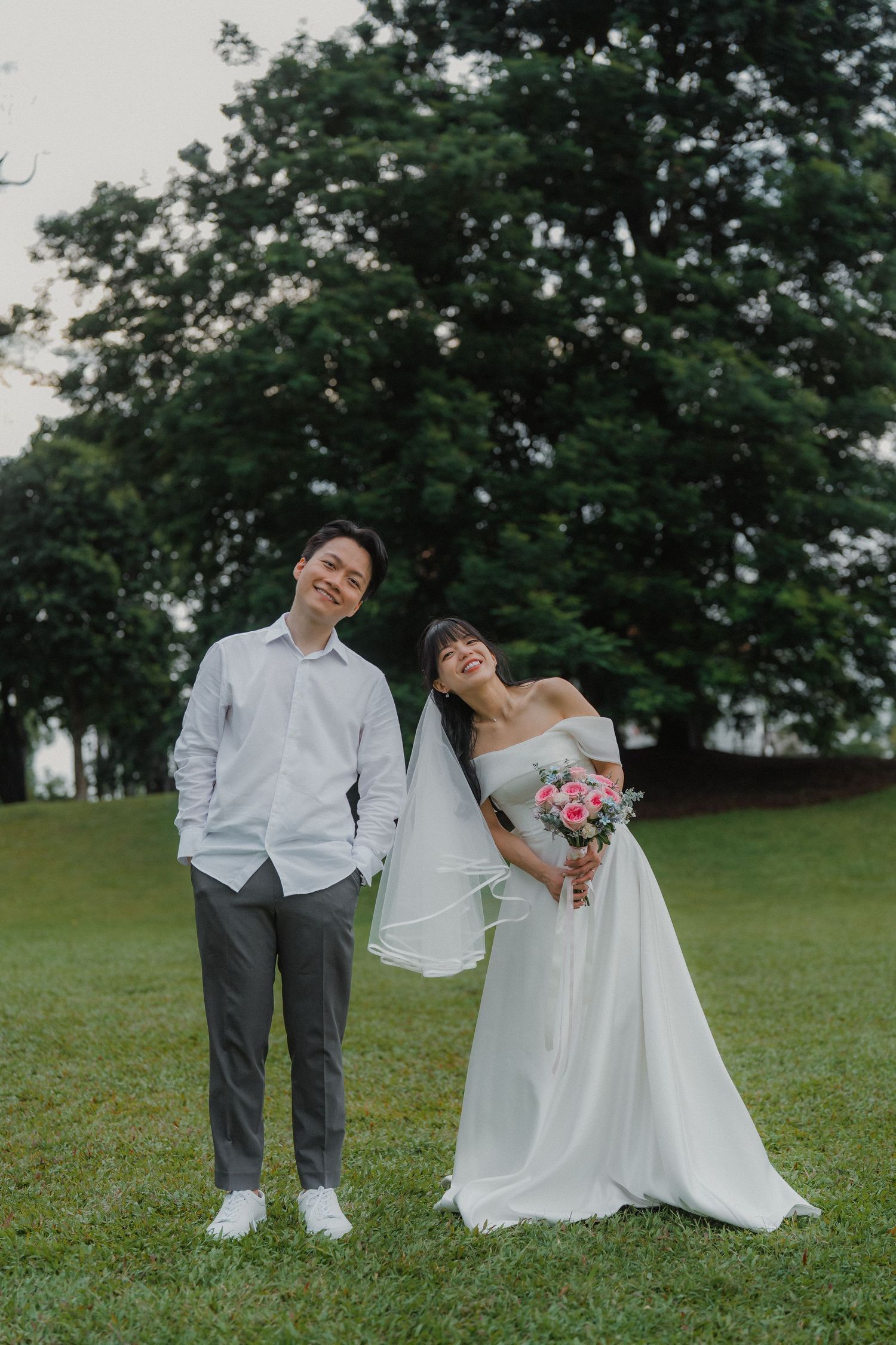 Wedding couple poses playfully on green lawn with tall trees in background.