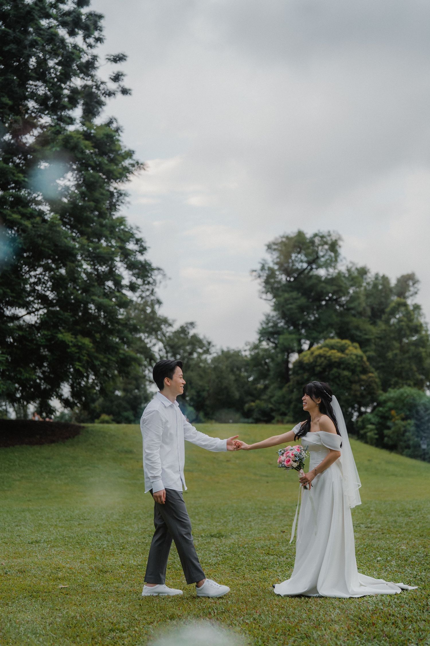 A wedding couple holds hands in a serene garden setting under cloudy skies.