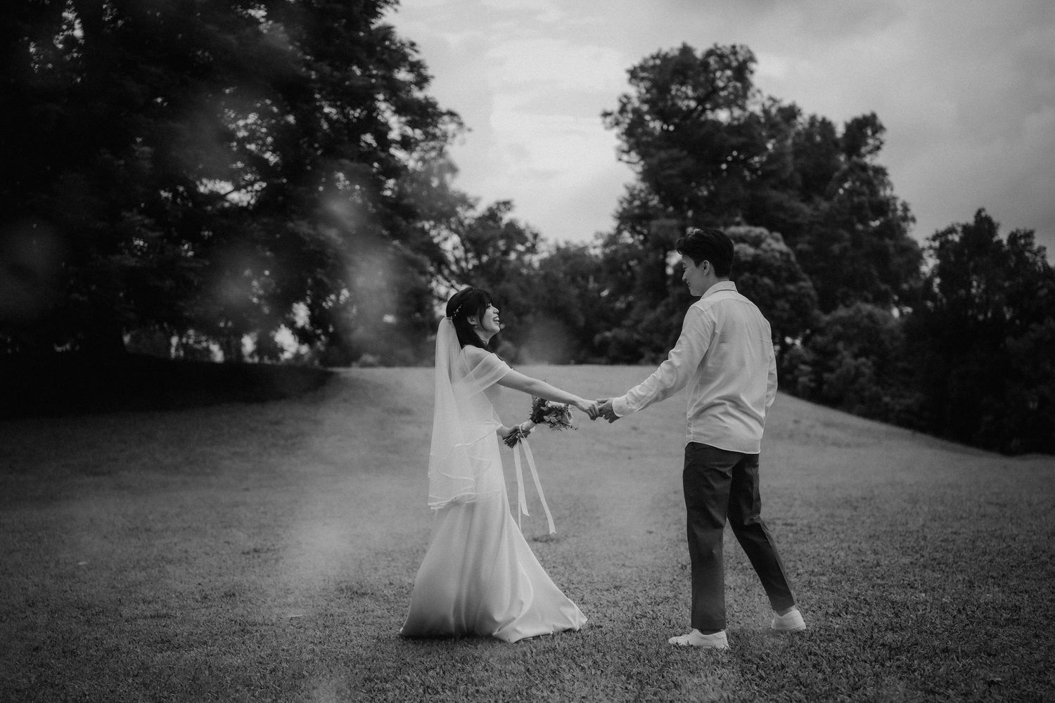 A black and white photo of a couple dancing playfully on a grassy lawn.