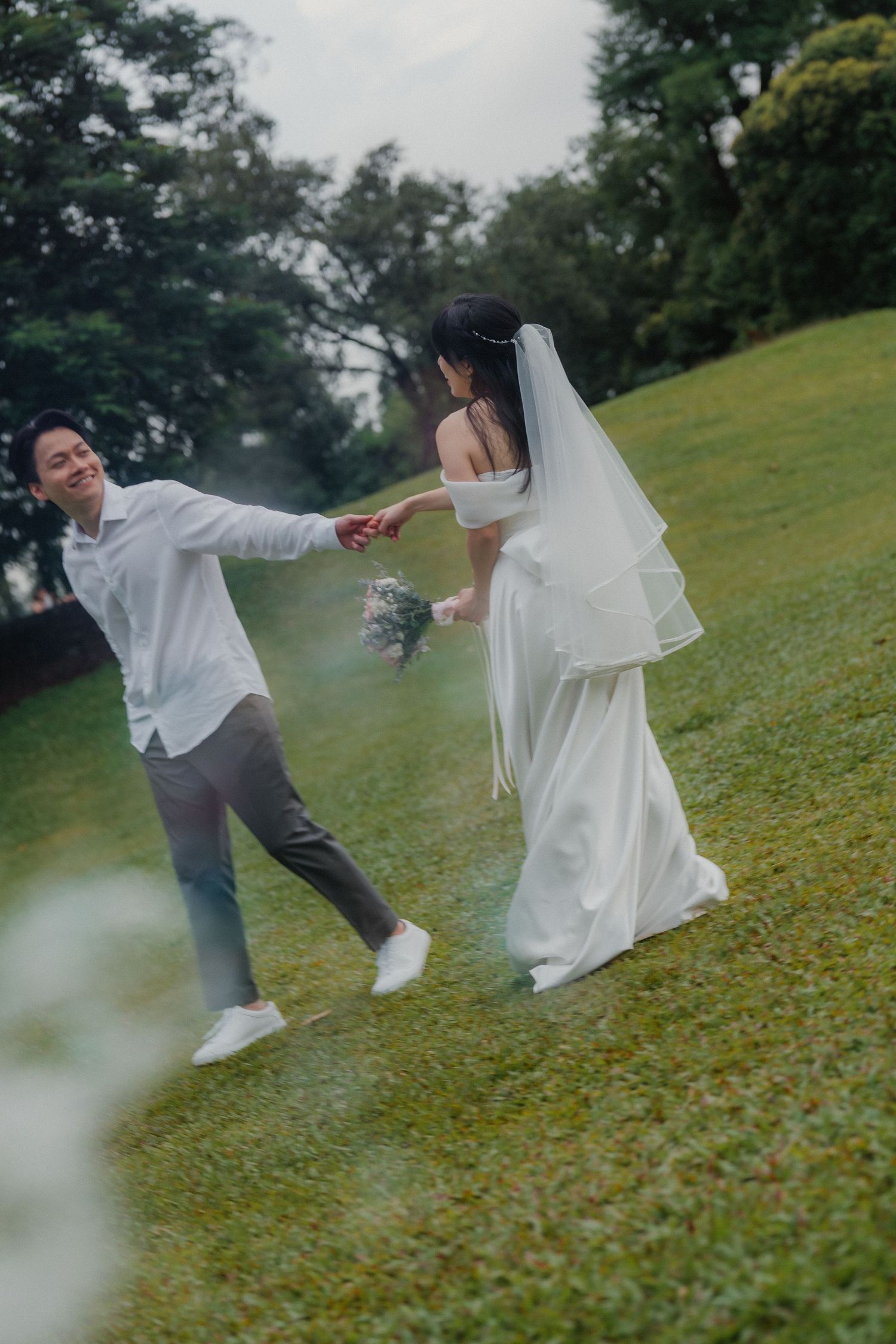 A playful wedding couple runs hand-in-hand across a grassy hillside.