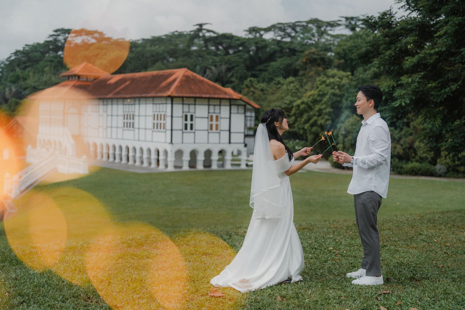 A playful wedding photo with lens flare as couple poses on grass with colonial building in background.