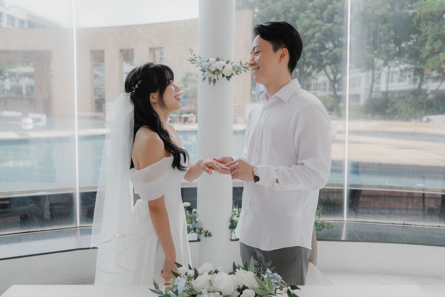 Couple exchanging rings during wedding ceremony in modern glass-walled venue.