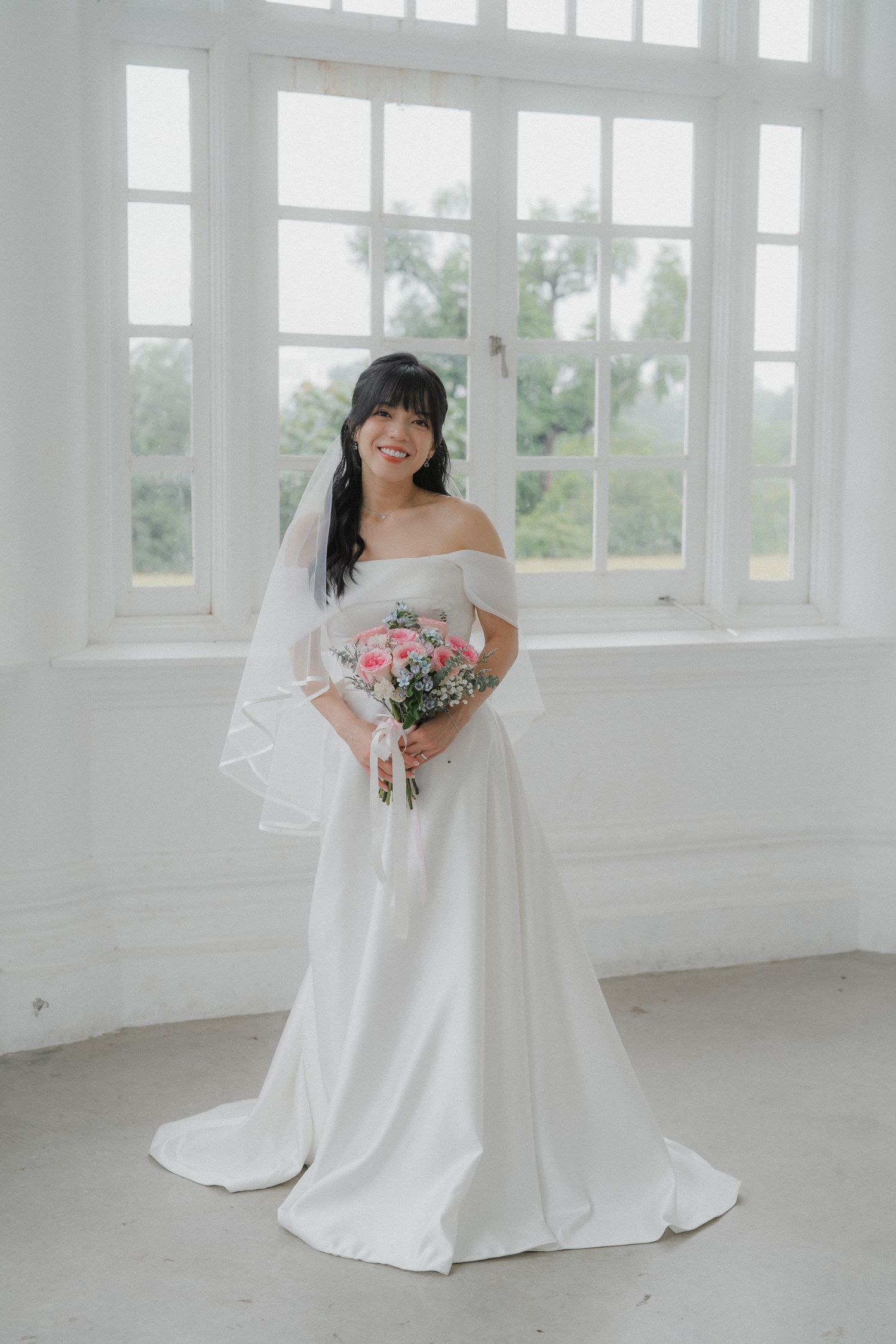 A bride in an off-shoulder wedding gown poses by bright windows holding a pink bouquet.
