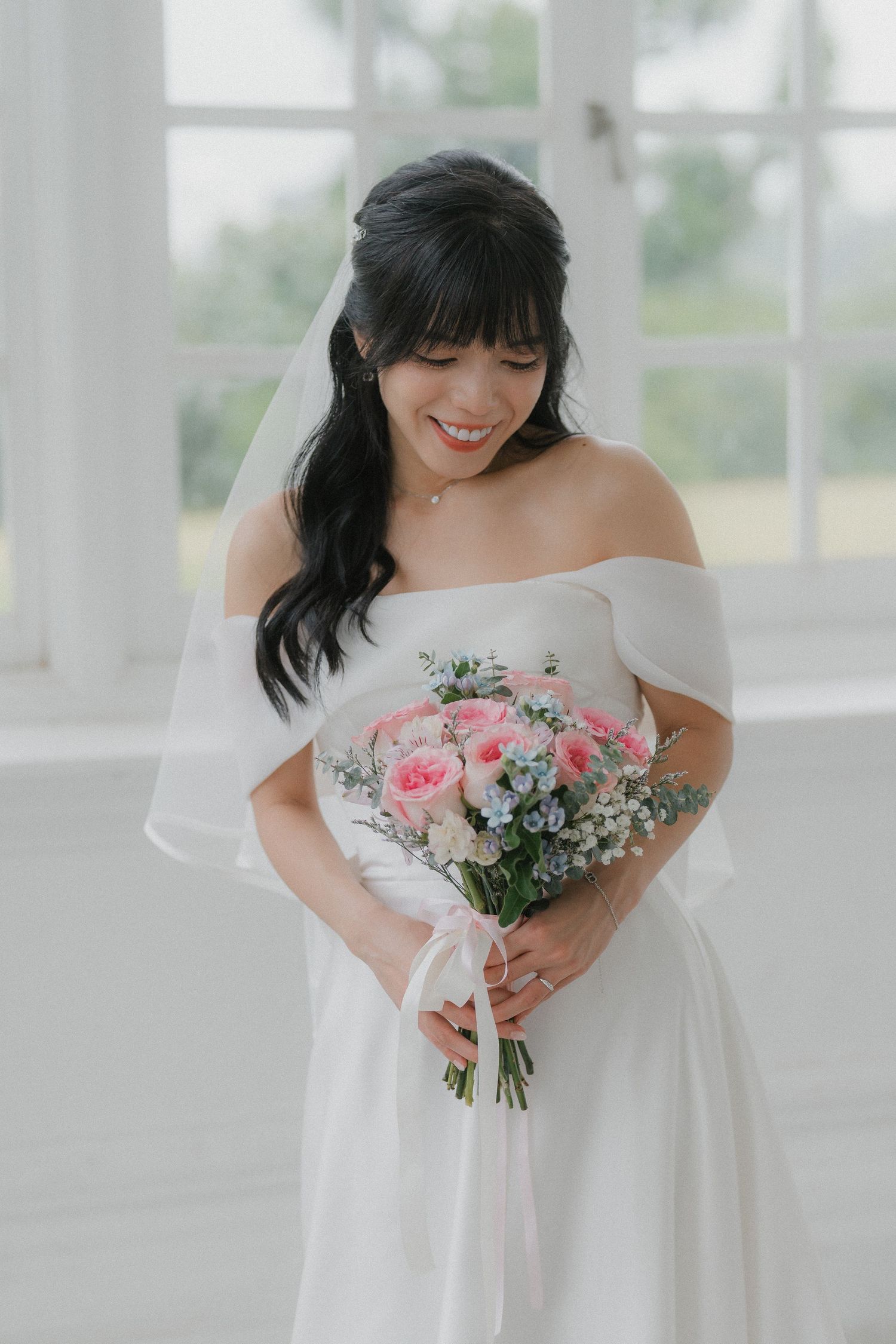 A joyful bride in an off-shoulder wedding gown holds a pink and white floral bouquet in a bright room with windows.