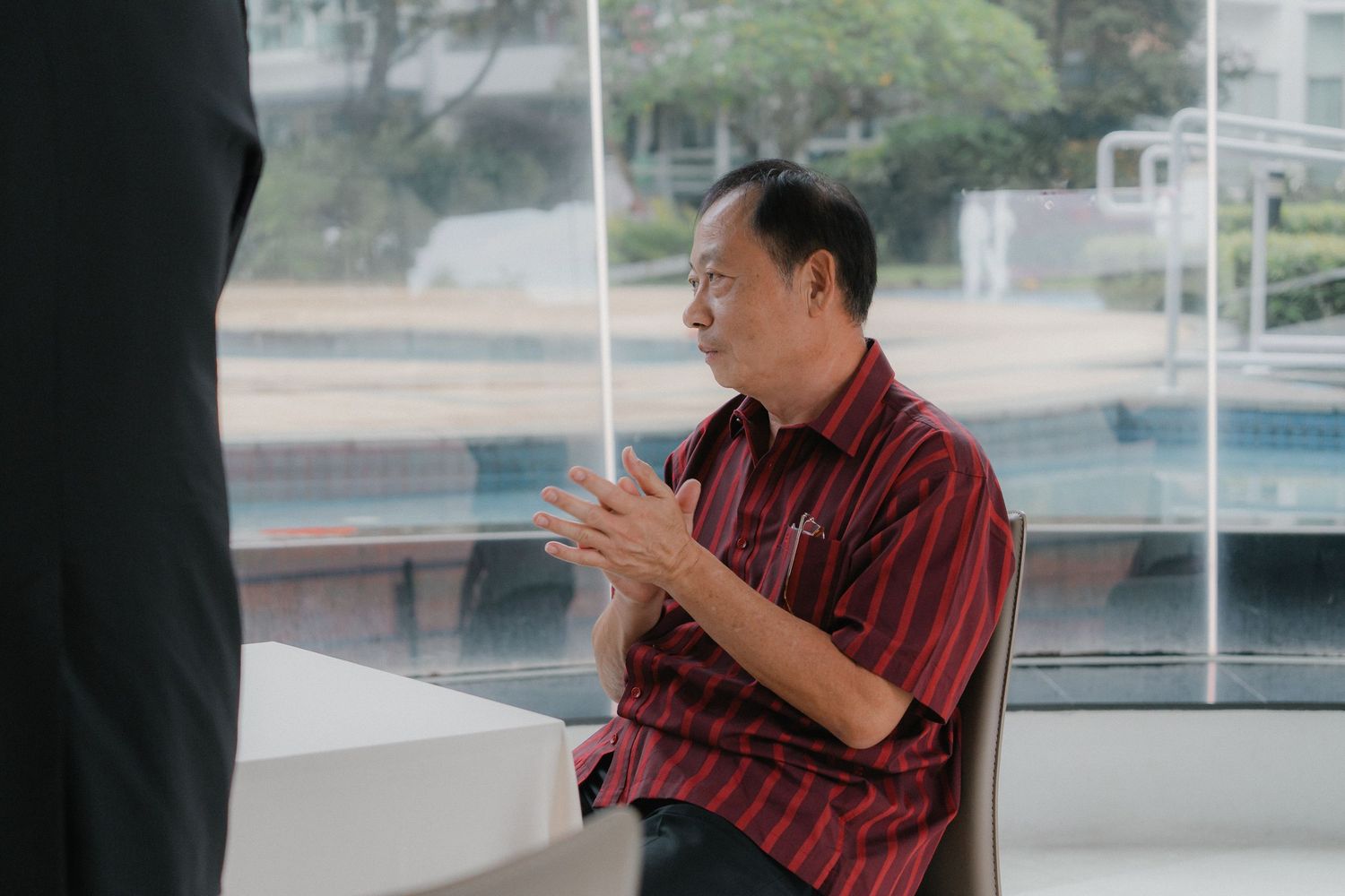 Person wearing maroon shirt sitting indoors near large windows in contemplative pose.