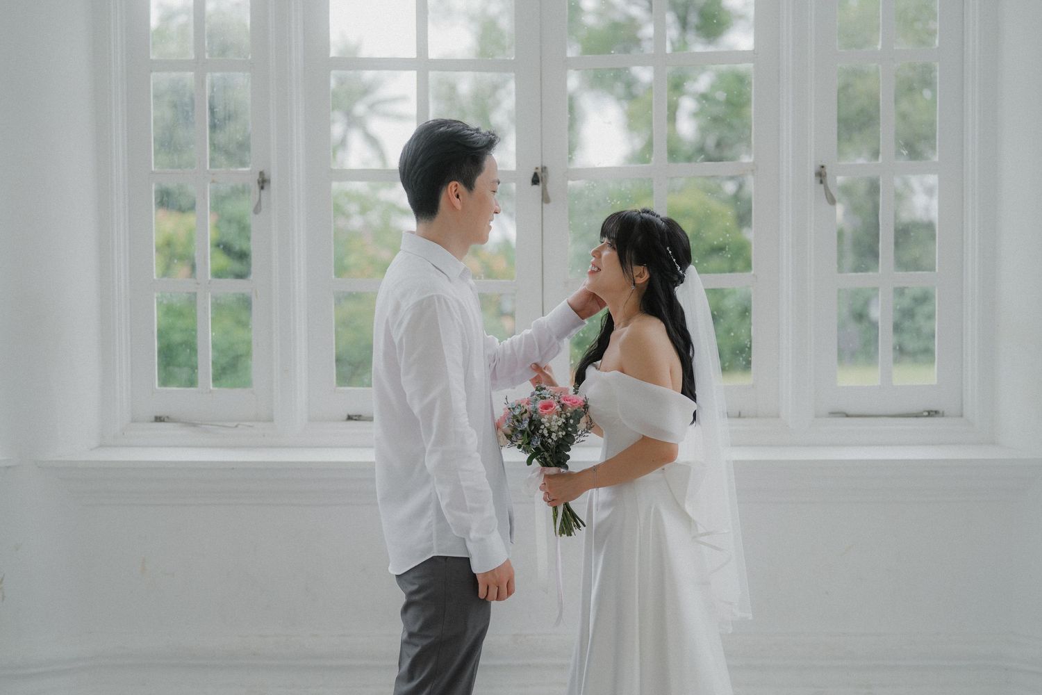 A couple in formal wedding attire shares an intimate moment in a white sunlit room.