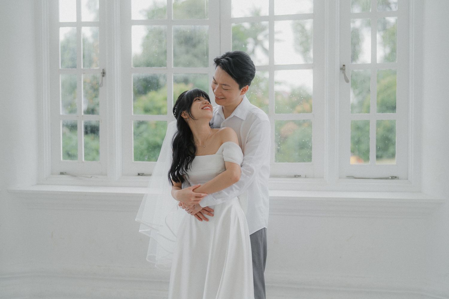 A romantic wedding couple embraces in a bright white room with large windows.