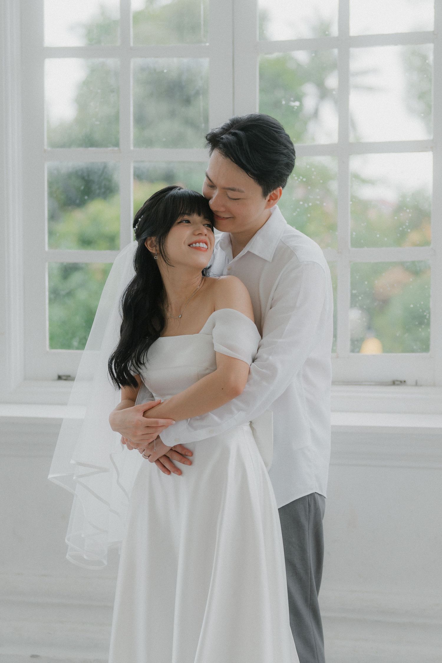 A romantic wedding portrait of a couple embracing by a bright window.