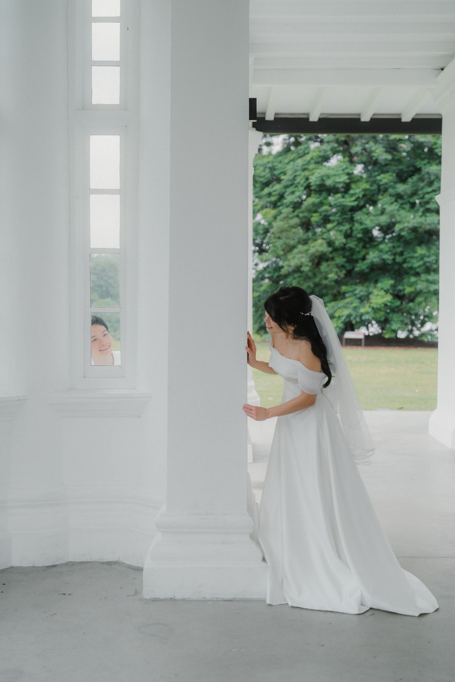 A bride in a white off-shoulder gown stands near a window in a bright white room.