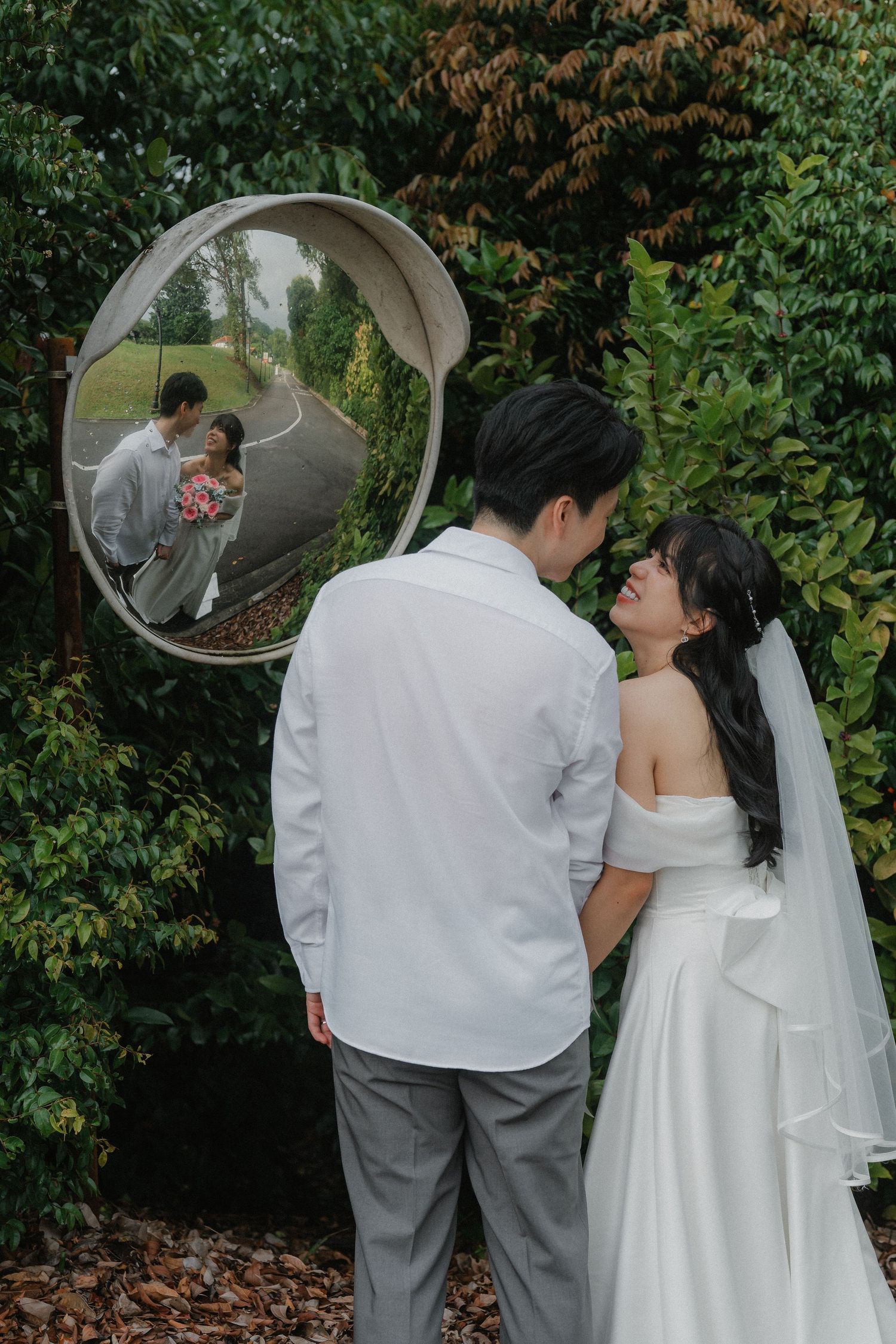 A creative wedding photo using a circular mirror reflection showing a couple in white and grey attire among garden foliage.