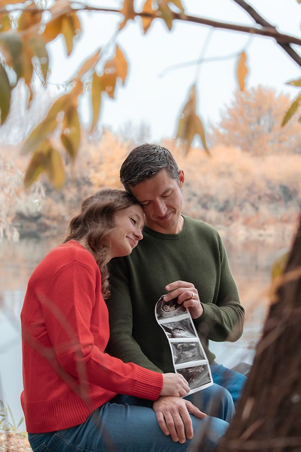 Couple sitting by the water during fall, holding a sonogram and smiling during a pregnancy announcement session in Tri-Cities WA