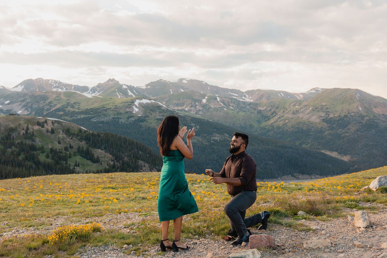 A couple shares a romantic moment on a mountaintop meadow with snow-capped peaks and wildflowers in the background.