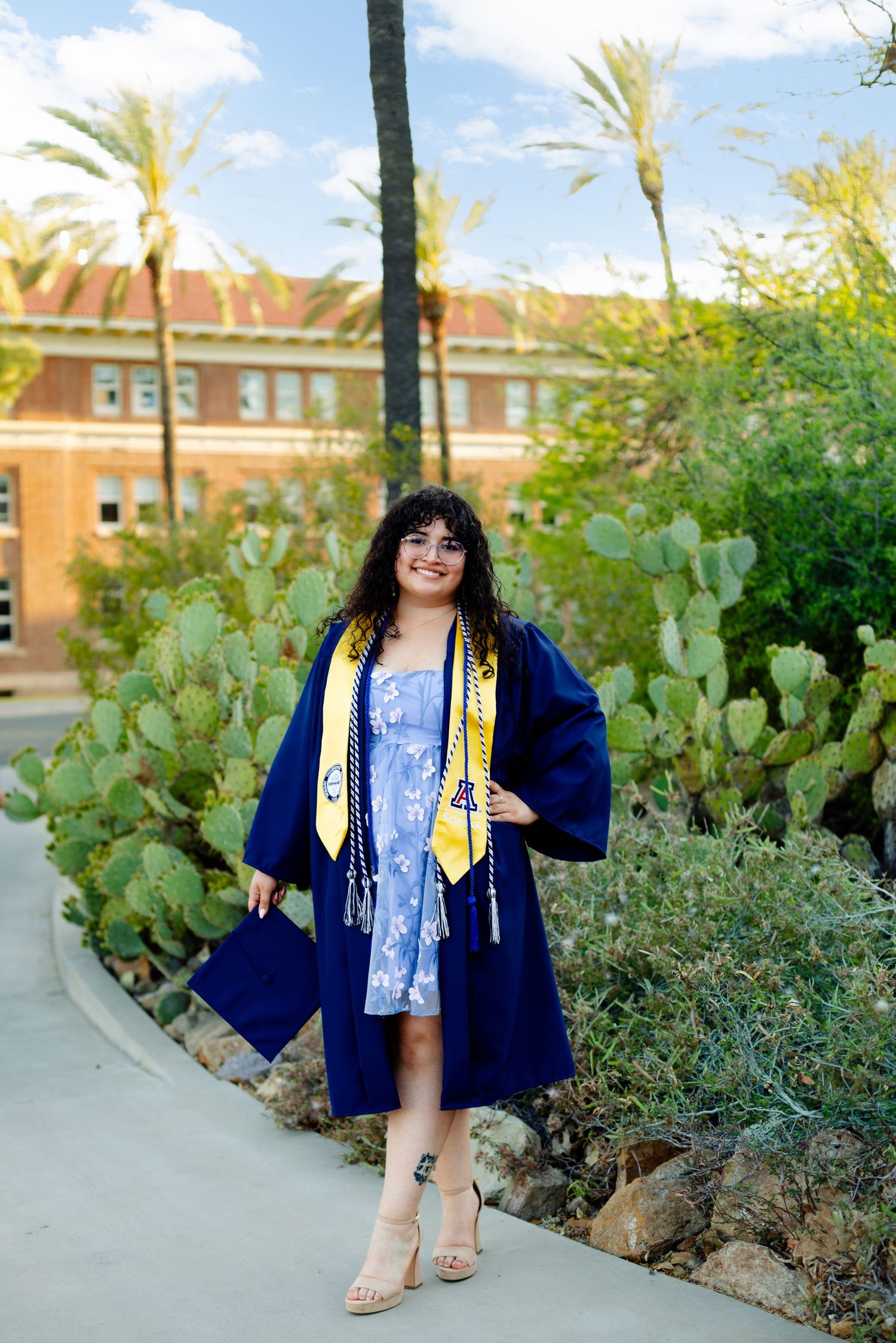 Graduate in blue cap and gown poses on campus walkway surrounded by desert landscaping.